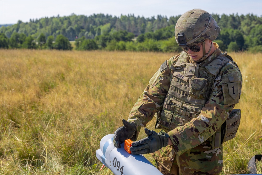 A U.S. Army soldier in full combat gear is handling what appears to be a small unmanned aerial vehicle (UAV) or drone component in an open field. The soldier, identified by visible name tape as 'JONES' with the rank of Specialist/Corporal, is manipulating an orange component on the gray UAV body. The setting appears to be a training exercise in a rural field environment with treeline in the background.