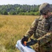 A U.S. Army soldier in full combat gear is handling what appears to be a small unmanned aerial vehicle (UAV) or drone component in an open field. The soldier, identified by visible name tape as 'JONES' with the rank of Specialist/Corporal, is manipulating an orange component on the gray UAV body. The setting appears to be a training exercise in a rural field environment with treeline in the background.