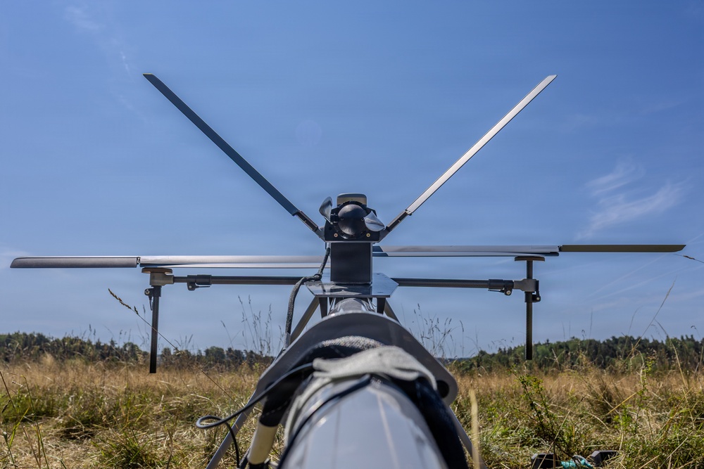 A close-up ground-level view of an unmanned aerial vehicle (UAV) or drone, photographed from behind looking toward its rotor assembly. The drone is stationary on dry grassland with a treeline visible in the background under a clear blue sky. The image captures the rotor blades, motor hub, landing gear struts, and fuselage in sharp detail.