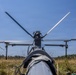 A close-up ground-level view of an unmanned aerial vehicle (UAV) or drone, photographed from behind looking toward its rotor assembly. The drone is stationary on dry grassland with a treeline visible in the background under a clear blue sky. The image captures the rotor blades, motor hub, landing gear struts, and fuselage in sharp detail.
