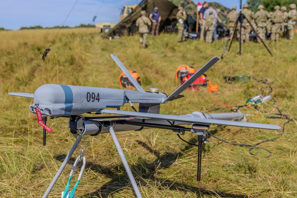 A military unmanned aerial vehicle (UAV) marked with the number '094' is displayed on a tripod stand in an open grassy field. The drone appears to be a fixed-wing reconnaissance or tactical UAV with a red 'Remove Before Flight' safety tag attached. In the background, military personnel in camouflage uniforms are gathered near equipment and a tent structure, suggesting a field exercise or demonstration.