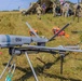 A military unmanned aerial vehicle (UAV) marked with the number '094' is displayed on a tripod stand in an open grassy field. The drone appears to be a fixed-wing reconnaissance or tactical UAV with a red 'Remove Before Flight' safety tag attached. In the background, military personnel in camouflage uniforms are gathered near equipment and a tent structure, suggesting a field exercise or demonstration.