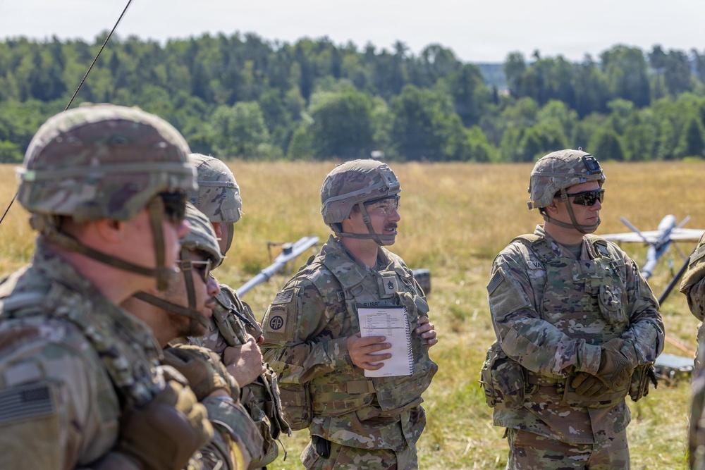 A group of U.S. Army soldiers in MultiCam/OCP camouflage uniforms and combat helmets are gathered in an open field during what appears to be a training briefing or after-action review. One soldier in the center is holding a notebook and appears to be speaking to the group. Military unmanned aerial vehicles (UAVs/drones) are visible in the background on the field.