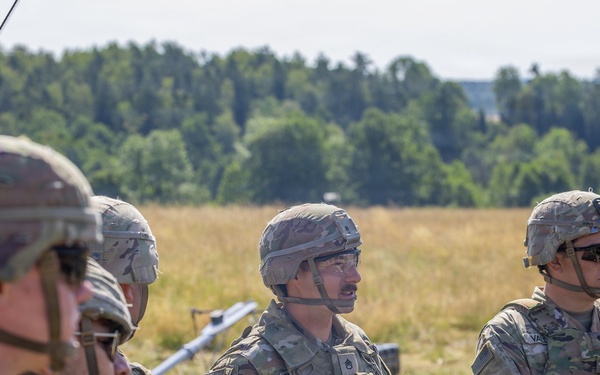 A group of U.S. Army soldiers in MultiCam/OCP camouflage uniforms and combat helmets are gathered in an open field during what appears to be a training briefing or after-action review. One soldier in the center is holding a notebook and appears to be speaking to the group. Military unmanned aerial vehicles (UAVs/drones) are visible in the background on the field.