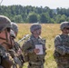 A group of U.S. Army soldiers in MultiCam/OCP camouflage uniforms and combat helmets are gathered in an open field during what appears to be a training briefing or after-action review. One soldier in the center is holding a notebook and appears to be speaking to the group. Military unmanned aerial vehicles (UAVs/drones) are visible in the background on the field.