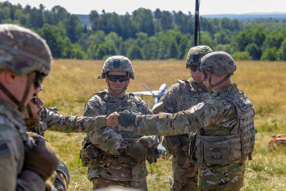 A group of U.S. Army soldiers in full combat gear are gathered in an open field, appearing to conduct a briefing or coordination activity. One soldier is pointing or gesturing toward others, suggesting instruction or mission planning. A small unmanned aerial vehicle (UAV/drone) is visible in the background on the field.