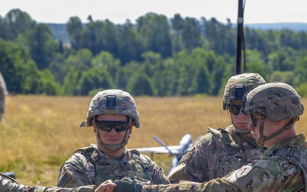 A group of U.S. Army soldiers in full combat gear are gathered in an open field, appearing to conduct a briefing or coordination activity. One soldier is pointing or gesturing toward others, suggesting instruction or mission planning. A small unmanned aerial vehicle (UAV/drone) is visible in the background on the field.