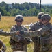 A group of U.S. Army soldiers in full combat gear are gathered in an open field, appearing to conduct a briefing or coordination activity. One soldier is pointing or gesturing toward others, suggesting instruction or mission planning. A small unmanned aerial vehicle (UAV/drone) is visible in the background on the field.