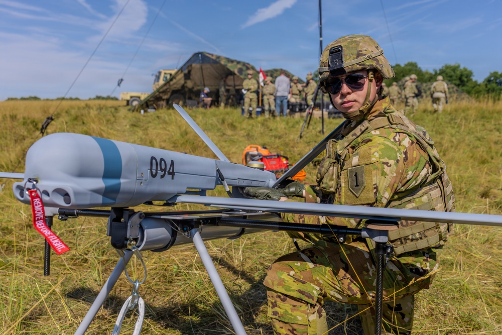 A U.S. Army soldier in OCP (Operational Camouflage Pattern) uniform is conducting pre-flight checks or maintenance on a small unmanned aerial vehicle (UAV) numbered '094', which bears a red 'Remove Before Flight' safety tag. The soldier wears a helmet with a mounted device and sunglasses, and displays a '1st Infantry Division' patch on the left shoulder. A forward operating base with military tents, vehicles, antennas, and additional personnel is visible in the background.
