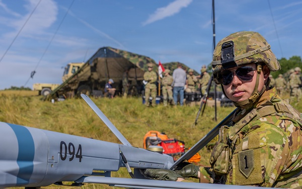 A U.S. Army soldier in OCP (Operational Camouflage Pattern) uniform is conducting pre-flight checks or maintenance on a small unmanned aerial vehicle (UAV) numbered '094', which bears a red 'Remove Before Flight' safety tag. The soldier wears a helmet with a mounted device and sunglasses, and displays a '1st Infantry Division' patch on the left shoulder. A forward operating base with military tents, vehicles, antennas, and additional personnel is visible in the background.