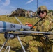 A U.S. Army soldier in OCP (Operational Camouflage Pattern) uniform is conducting pre-flight checks or maintenance on a small unmanned aerial vehicle (UAV) numbered '094', which bears a red 'Remove Before Flight' safety tag. The soldier wears a helmet with a mounted device and sunglasses, and displays a '1st Infantry Division' patch on the left shoulder. A forward operating base with military tents, vehicles, antennas, and additional personnel is visible in the background.
