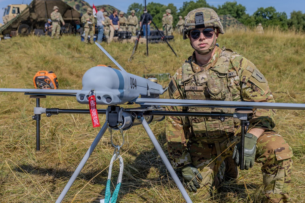 A U.S. Army soldier identified by name tape as 'Jones' kneels beside a fixed-wing unmanned aerial vehicle (UAV/drone) marked '094' on an open grassy field. The soldier is in full combat uniform with OCP camouflage, helmet, sunglasses, and tactical gear. In the background, multiple other soldiers and military equipment including a vehicle and tent are visible, suggesting a field exercise or demonstration.