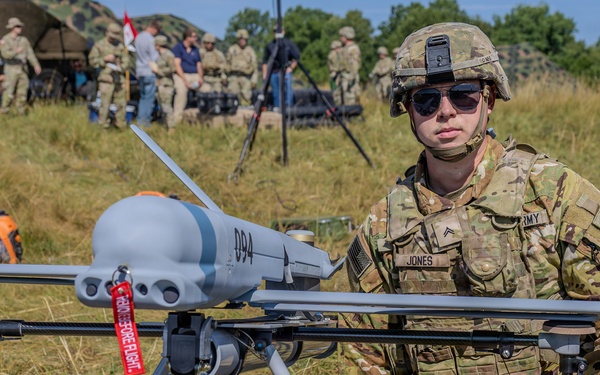 A U.S. Army soldier identified by name tape as 'Jones' kneels beside a fixed-wing unmanned aerial vehicle (UAV/drone) marked '094' on an open grassy field. The soldier is in full combat uniform with OCP camouflage, helmet, sunglasses, and tactical gear. In the background, multiple other soldiers and military equipment including a vehicle and tent are visible, suggesting a field exercise or demonstration.