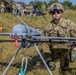A U.S. Army soldier identified by name tape as 'Jones' kneels beside a fixed-wing unmanned aerial vehicle (UAV/drone) marked '094' on an open grassy field. The soldier is in full combat uniform with OCP camouflage, helmet, sunglasses, and tactical gear. In the background, multiple other soldiers and military equipment including a vehicle and tent are visible, suggesting a field exercise or demonstration.