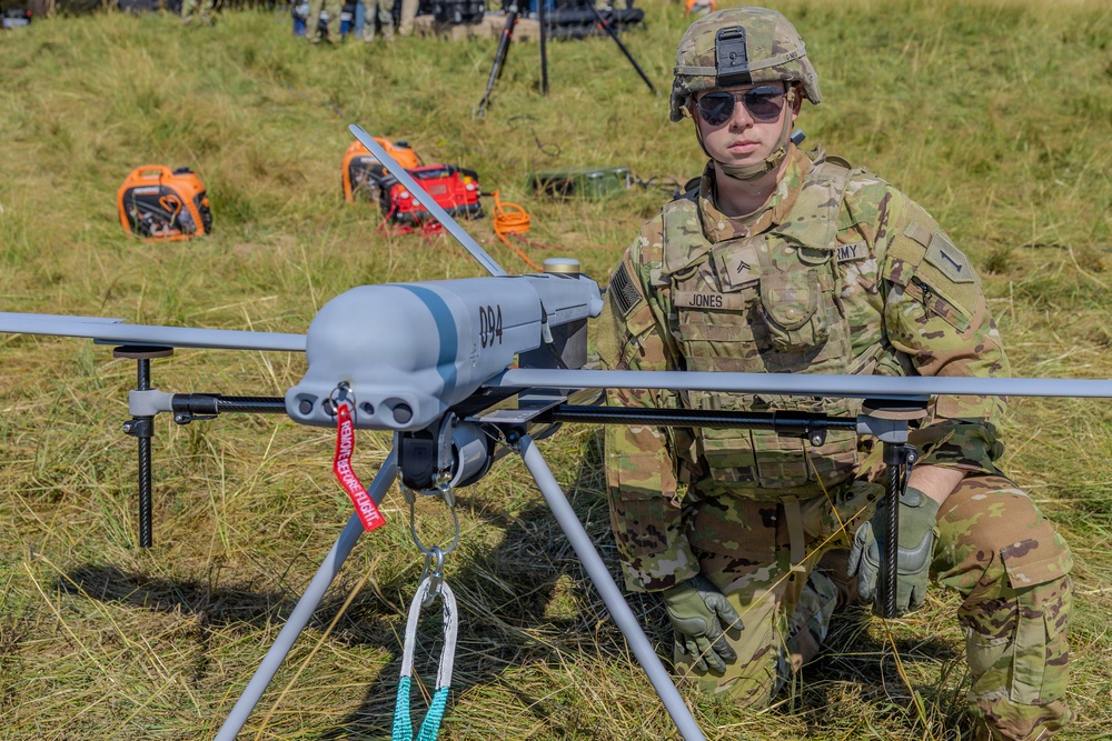 A U.S. Army soldier identified by name tape as 'JONES' with the rank of Corporal kneels beside a fixed-wing military unmanned aerial vehicle (UAV/drone) marked with the number '894' on a grassy field. The soldier is wearing MultiCam (OCP) camouflage uniform, combat helmet, sunglasses, and tactical gloves. The scene appears to be a military demonstration or pre-flight preparation exercise.