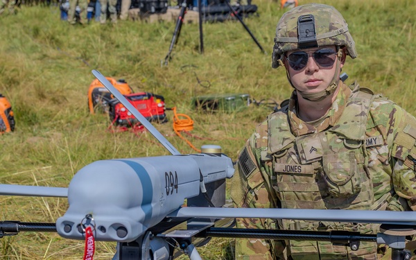 A U.S. Army soldier identified by name tape as 'JONES' with the rank of Corporal kneels beside a fixed-wing military unmanned aerial vehicle (UAV/drone) marked with the number '894' on a grassy field. The soldier is wearing MultiCam (OCP) camouflage uniform, combat helmet, sunglasses, and tactical gloves. The scene appears to be a military demonstration or pre-flight preparation exercise.