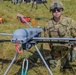 A U.S. Army soldier identified by name tape as 'JONES' with the rank of Corporal kneels beside a fixed-wing military unmanned aerial vehicle (UAV/drone) marked with the number '894' on a grassy field. The soldier is wearing MultiCam (OCP) camouflage uniform, combat helmet, sunglasses, and tactical gloves. The scene appears to be a military demonstration or pre-flight preparation exercise.