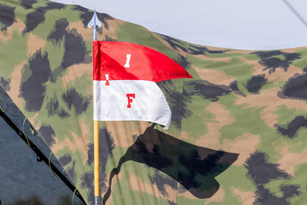 The image shows a military-style pennant flag mounted on a yellow pole with a spear-tip finial, displayed against a camouflage tent or tarpaulin backdrop. The flag is divided horizontally into red (top) and white (bottom) halves, bearing the markings '1' on the red section and 'F' on the white section. The setting appears to be a military exercise, reenactment, or field demonstration.