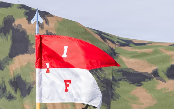 The image shows a military-style pennant flag mounted on a yellow pole with a spear-tip finial, displayed against a camouflage tent or tarpaulin backdrop. The flag is divided horizontally into red (top) and white (bottom) halves, bearing the markings '1' on the red section and 'F' on the white section. The setting appears to be a military exercise, reenactment, or field demonstration.