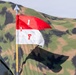 The image shows a military-style pennant flag mounted on a yellow pole with a spear-tip finial, displayed against a camouflage tent or tarpaulin backdrop. The flag is divided horizontally into red (top) and white (bottom) halves, bearing the markings '1' on the red section and 'F' on the white section. The setting appears to be a military exercise, reenactment, or field demonstration.