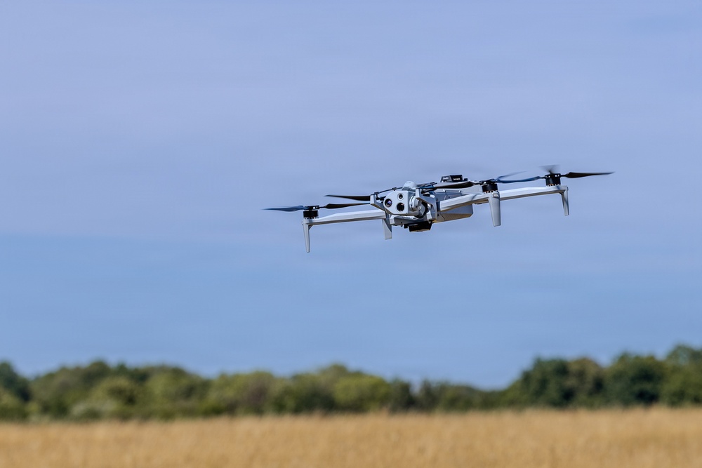 A consumer-grade quadcopter drone is captured mid-flight against a blue sky backdrop. The drone appears to be hovering or flying at low altitude over an open field with dry golden grass and a treeline visible in the background. The image is sharp and well-composed, with the drone as the clear focal subject.