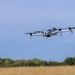 A consumer-grade quadcopter drone is captured mid-flight against a blue sky backdrop. The drone appears to be hovering or flying at low altitude over an open field with dry golden grass and a treeline visible in the background. The image is sharp and well-composed, with the drone as the clear focal subject.