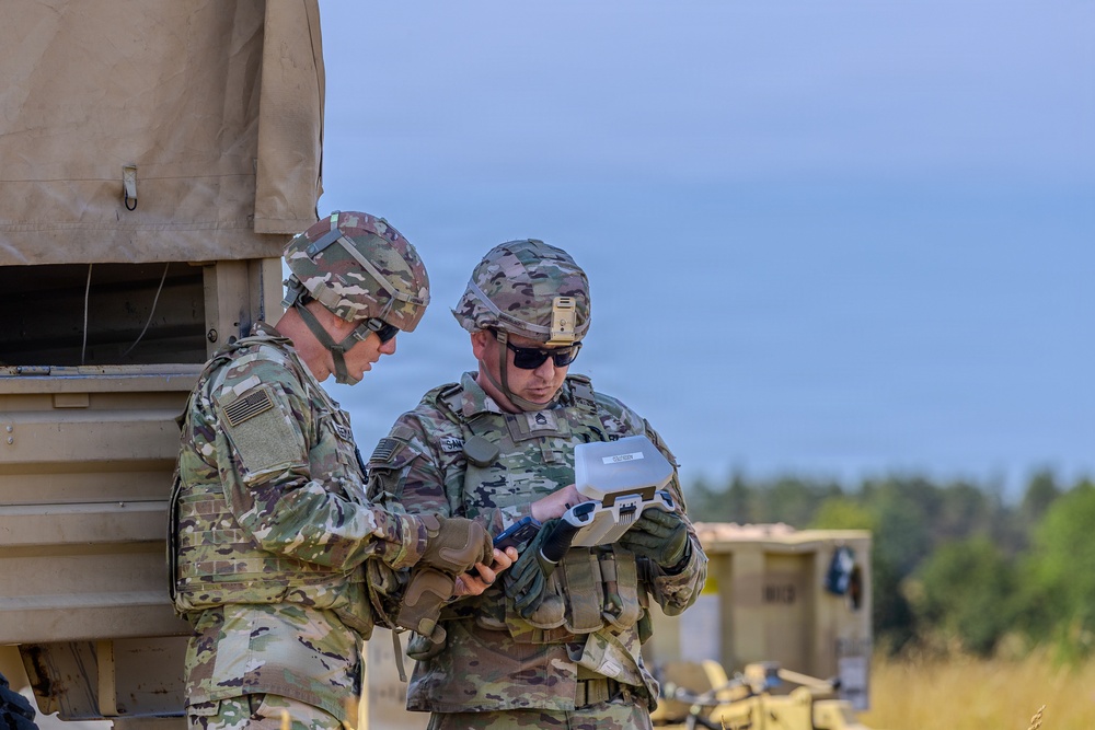 Two military personnel in US Army MultiCam/OCP camouflage uniforms are examining a device or document together near a military vehicle. Both soldiers are wearing combat helmets, tactical vests, and sunglasses, suggesting an active field exercise or operational environment. Additional military vehicles are visible in the background.