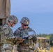 Two military personnel in US Army MultiCam/OCP camouflage uniforms are examining a device or document together near a military vehicle. Both soldiers are wearing combat helmets, tactical vests, and sunglasses, suggesting an active field exercise or operational environment. Additional military vehicles are visible in the background.