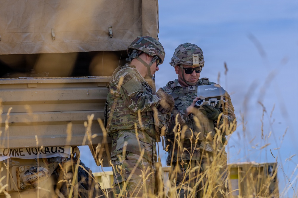 Two soldiers in U.S. Army Operational Camouflage Pattern (OCP) uniforms are examining a small device, likely a tablet or handheld controller, near a military vehicle or equipment structure. The text 'KOLONNE VORAUS' (German for 'convoy ahead') is partially visible on the vehicle, suggesting a NATO or joint exercise environment. The setting appears to be an open field during daylight hours.
