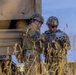 Two soldiers in U.S. Army Operational Camouflage Pattern (OCP) uniforms are examining a small device, likely a tablet or handheld controller, near a military vehicle or equipment structure. The text 'KOLONNE VORAUS' (German for 'convoy ahead') is partially visible on the vehicle, suggesting a NATO or joint exercise environment. The setting appears to be an open field during daylight hours.