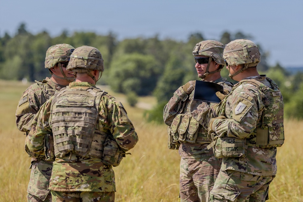 Four U.S. Army soldiers in full combat gear are gathered in an open field, appearing to conduct a briefing or discussion. They are wearing Operational Camouflage Pattern (OCP) uniforms with helmets and body armor. The setting appears to be a training area or field exercise location.