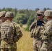 Four U.S. Army soldiers in full combat gear are gathered in an open field, appearing to conduct a briefing or discussion. They are wearing Operational Camouflage Pattern (OCP) uniforms with helmets and body armor. The setting appears to be a training area or field exercise location.