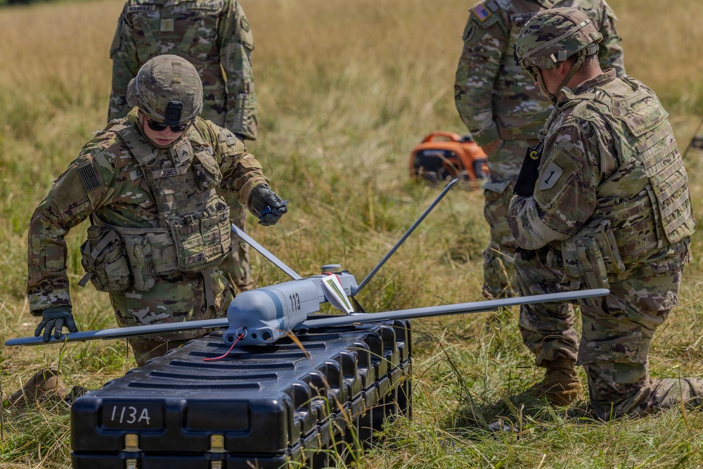 U.S. Army soldiers in MultiCam/OCP camouflage uniforms are preparing a fixed-wing UAV (unmanned aerial vehicle) in an open grassy field. The drone, marked with the number '113', is resting on a hard-shell transport case labeled '113A'. The soldiers appear to be conducting pre-launch assembly or inspection procedures.