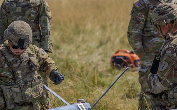 U.S. Army soldiers in MultiCam/OCP camouflage uniforms are preparing a fixed-wing UAV (unmanned aerial vehicle) in an open grassy field. The drone, marked with the number '113', is resting on a hard-shell transport case labeled '113A'. The soldiers appear to be conducting pre-launch assembly or inspection procedures.