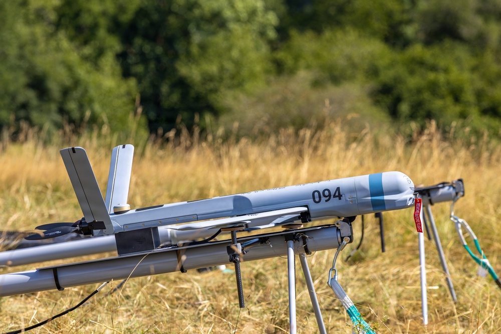 The image shows a fixed-wing unmanned aerial vehicle (UAV) mounted on a launch rail or catapult system in an outdoor field setting. The aircraft is light gray with the number '094' marked on its fuselage and features a rear-mounted pusher propeller configuration. The UAV appears to be prepared for launch, positioned on a tripod-supported rail system.