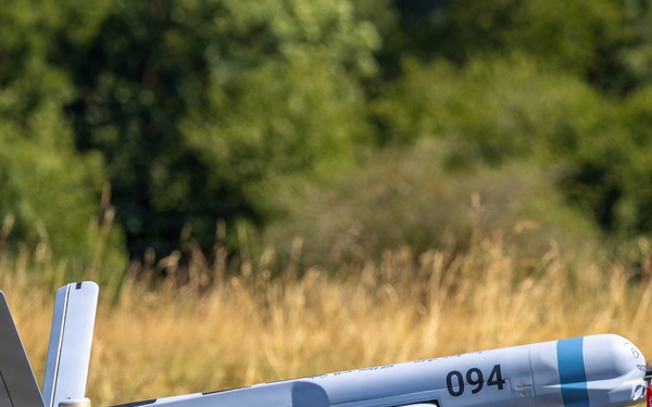 The image shows a fixed-wing unmanned aerial vehicle (UAV) mounted on a launch rail or catapult system in an outdoor field setting. The aircraft is light gray with the number '094' marked on its fuselage and features a rear-mounted pusher propeller configuration. The UAV appears to be prepared for launch, positioned on a tripod-supported rail system.