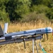 The image shows a fixed-wing unmanned aerial vehicle (UAV) mounted on a launch rail or catapult system in an outdoor field setting. The aircraft is light gray with the number '094' marked on its fuselage and features a rear-mounted pusher propeller configuration. The UAV appears to be prepared for launch, positioned on a tripod-supported rail system.
