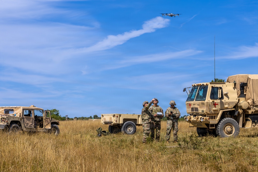 Three military personnel in camouflage uniforms are operating a small quadcopter drone in an open field. They are surrounded by military vehicles including a Humvee, a trailer, and a large tactical truck marked with a red cross indicating medical designation. The scene appears to be a training or field exercise conducted in a dry grassland environment.