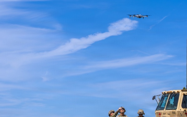 Three military personnel in camouflage uniforms are operating a small quadcopter drone in an open field. They are surrounded by military vehicles including a Humvee, a trailer, and a large tactical truck marked with a red cross indicating medical designation. The scene appears to be a training or field exercise conducted in a dry grassland environment.