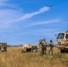 Three military personnel in camouflage uniforms are operating a small quadcopter drone in an open field. They are surrounded by military vehicles including a Humvee, a trailer, and a large tactical truck marked with a red cross indicating medical designation. The scene appears to be a training or field exercise conducted in a dry grassland environment.
