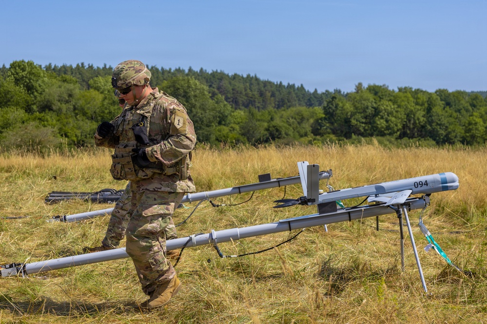 A U.S. Army soldier in MultiCam/OCP camouflage uniform is operating near a UAV (unmanned aerial vehicle) launch rail system in an open field. The soldier appears to be preparing or checking equipment associated with what appears to be a small fixed-wing drone on a catapult/rail launcher marked '094'. The setting is a dry grassy field with a tree line in the background under clear blue skies.