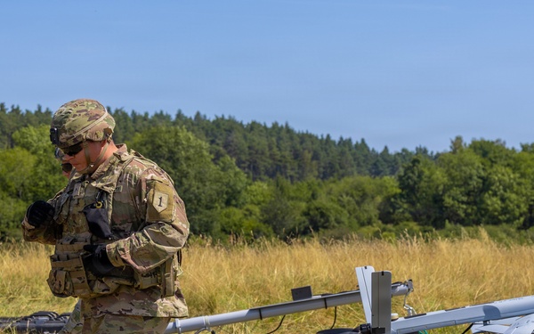 A U.S. Army soldier in MultiCam/OCP camouflage uniform is operating near a UAV (unmanned aerial vehicle) launch rail system in an open field. The soldier appears to be preparing or checking equipment associated with what appears to be a small fixed-wing drone on a catapult/rail launcher marked '094'. The setting is a dry grassy field with a tree line in the background under clear blue skies.