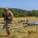 A U.S. Army soldier in MultiCam/OCP camouflage uniform is operating near a UAV (unmanned aerial vehicle) launch rail system in an open field. The soldier appears to be preparing or checking equipment associated with what appears to be a small fixed-wing drone on a catapult/rail launcher marked '094'. The setting is a dry grassy field with a tree line in the background under clear blue skies.