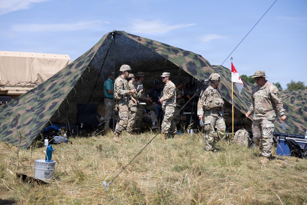 A group of military personnel in camouflage uniforms are gathered around a large camouflage-patterned field tent in an outdoor grassy area. Several soldiers appear to be engaged in briefings or coordination activities near the tent entrance. A military pennant flag on a pole is visible to the right of the tent.