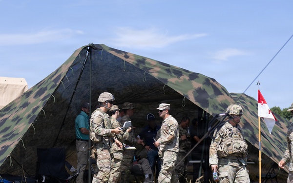 A group of military personnel in camouflage uniforms are gathered around a large camouflage-patterned field tent in an outdoor grassy area. Several soldiers appear to be engaged in briefings or coordination activities near the tent entrance. A military pennant flag on a pole is visible to the right of the tent.