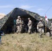 A group of military personnel in camouflage uniforms are gathered around a large camouflage-patterned field tent in an outdoor grassy area. Several soldiers appear to be engaged in briefings or coordination activities near the tent entrance. A military pennant flag on a pole is visible to the right of the tent.