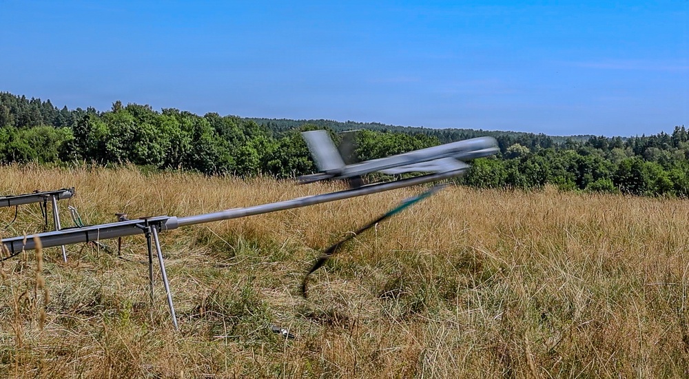 A fixed-wing UAV (unmanned aerial vehicle) is being launched from a catapult rail system in an open grassy field. The drone appears to be in motion, captured mid-launch with visible motion blur on its wings and propeller. The setting is a rural or semi-rural area surrounded by dense tree lines under clear blue skies.