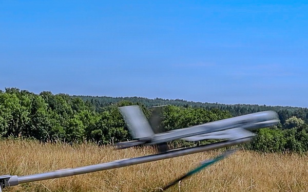 A fixed-wing UAV (unmanned aerial vehicle) is being launched from a catapult rail system in an open grassy field. The drone appears to be in motion, captured mid-launch with visible motion blur on its wings and propeller. The setting is a rural or semi-rural area surrounded by dense tree lines under clear blue skies.