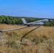 A fixed-wing UAV (unmanned aerial vehicle) is being launched from a catapult rail system in an open grassy field. The drone appears to be in motion, captured mid-launch with visible motion blur on its wings and propeller. The setting is a rural or semi-rural area surrounded by dense tree lines under clear blue skies.