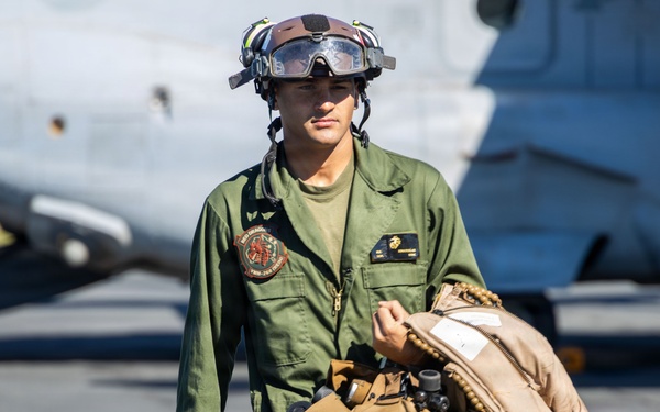A U.S. Marine Corps aviator or aircrew member is walking across a flight line tarmac in olive green flight suit. The individual is carrying flight gear including a helmet, survival vest, and what appears to be a flight jacket or parachute pack. A military aircraft is visible in the background.